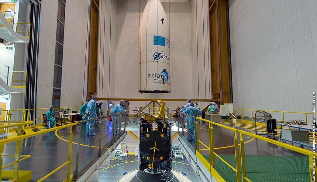 The nose cone of a rocket hangs in its factory with workers gathered around