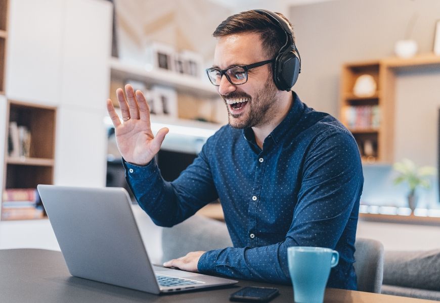 Man on a virtual call with a headset and waving to the laptop screen.